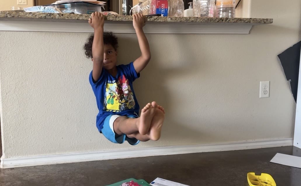 A young boy hanging from the kitchen counter in a tuck hold, habits learned by watching his parents